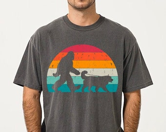 May include: A dark grey t-shirt featuring a vintage-style graphic of a Bigfoot walking a dog in front of a sunset. The graphic is in shades of orange, yellow, red, blue, and green.