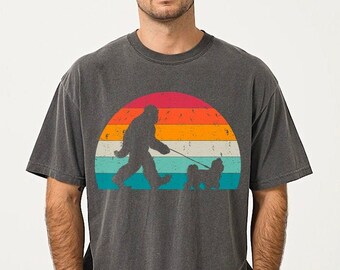 May include: A dark grey t-shirt featuring a retro-style graphic of a Bigfoot walking a small dog in front of a sunset. The sunset is a gradient of orange, yellow, blue, and green.