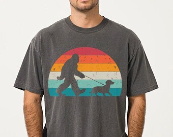 May include: A dark grey t-shirt featuring a vintage-style graphic of a Bigfoot walking a dachshund in front of a sunset.