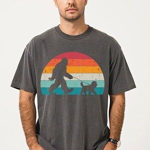 May include: A dark gray t-shirt with a vintage-style graphic of a Bigfoot walking a dog in front of a sunset. The graphic is in shades of orange, yellow, blue, and green.
