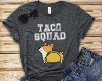 May include: A dark gray t-shirt with the text "Taco Squad" in white lettering. A cartoon corgi dog wearing a taco is printed on the shirt.