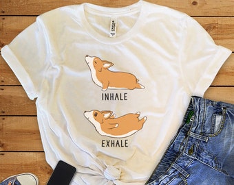 May include: White t-shirt with a graphic of a corgi dog in two poses, one with its belly up and the other with its belly down. The text "inhale" is above the first pose and "exhale" is below the second pose.