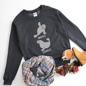 May include: A dark gray sweatshirt with a graphic of two manatees in a yoga pose. The text "inhale" is above the top manatee and "exhale" is below the bottom manatee.