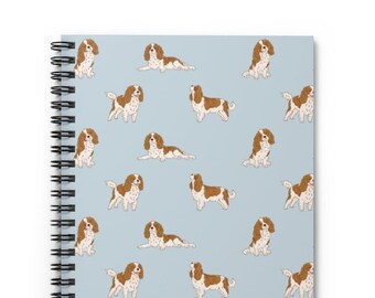May include: A blue spiral notebook with a repeating pattern of Cavalier King Charles Spaniel dogs. The dogs are brown and white and are in various poses.