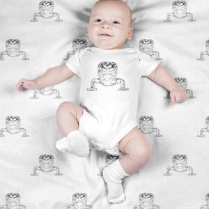 May include: A white baby onesie with a black and white gecko print. The onesie is on a white blanket with a repeating gecko pattern.