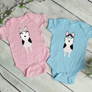 May include: Two baby onesies, one pink and one blue, with a black and white husky dog illustration. The pink onesie has a floral crown on the dog's head, and the blue onesie has the dog wearing glasses.