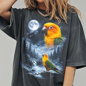 May include: Dark gray t-shirt featuring a vibrant graphic of two yellow and green parrots against a night scene with a full moon, waterfall, and forest. The shirt has a vintage, washed look.