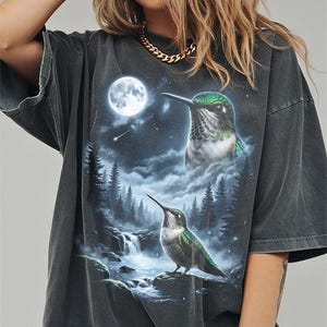 May include: Dark grey t-shirt with a detailed graphic of two hummingbirds, a full moon, and a waterfall scene. The birds have green and white feathers. The background includes a starry night sky and a forest.