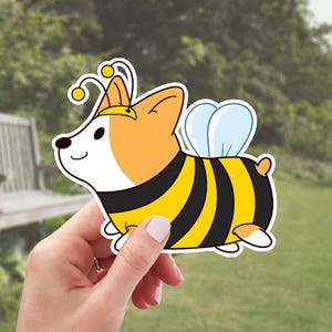 May include: A cartoon sticker of a corgi dressed as a bee. The corgi is wearing a yellow and black striped bee costume with wings and an antenna. The corgi is smiling and has a white face.