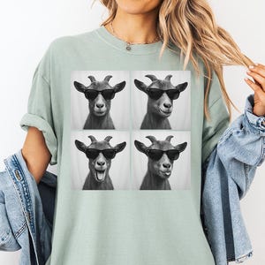 May include: A light green t-shirt featuring a black and white image of four goats wearing sunglasses. The goats have different facial expressions. The shirt is a relaxed fit.
