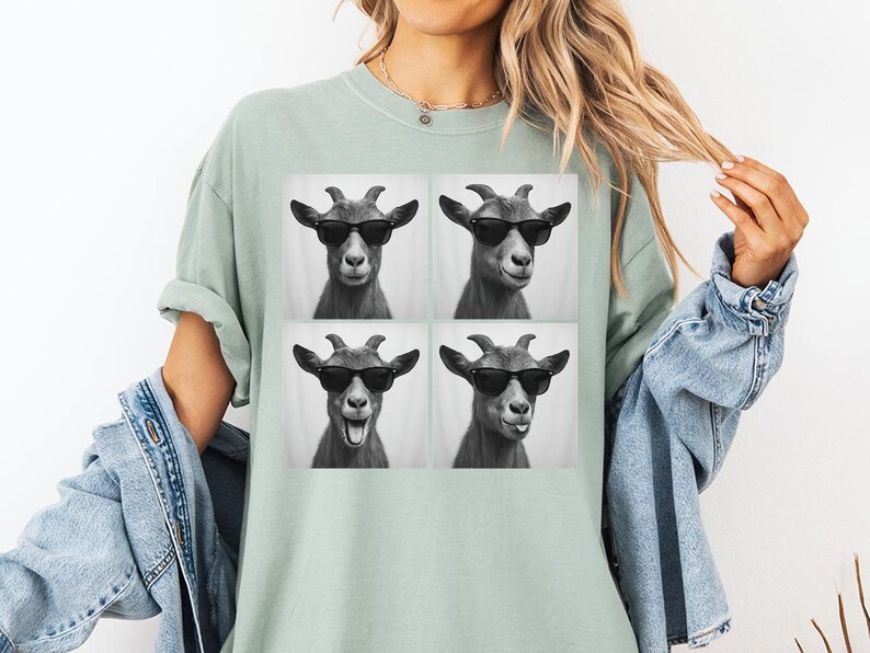 May include: A light green t-shirt featuring a black and white image of four goats wearing sunglasses. The goats have different facial expressions. The shirt is a relaxed fit.