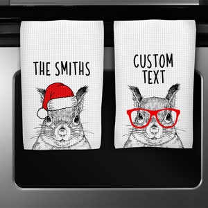 May include: Two white kitchen towels with a waffle weave texture. One towel features a squirrel wearing a Santa hat, the text "THE SMITHS" is above the squirrel. The other towel has a squirrel wearing red glasses, with the text "CUSTOM TEXT".