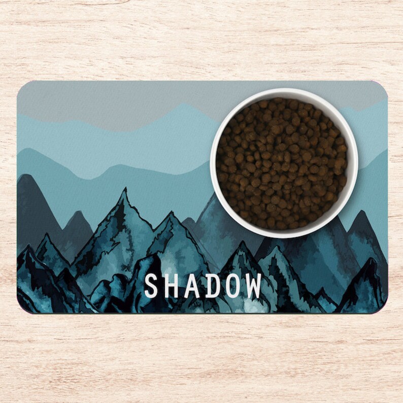 May include: A blue and gray mountain-themed pet food mat with the name "SHADOW" printed on it. A white bowl filled with brown dog food is on the mat.