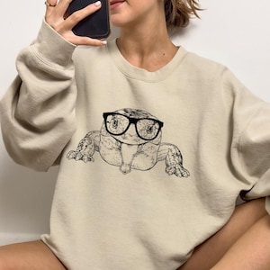 May include: A beige sweatshirt with a black and white illustration of a lizard wearing glasses.