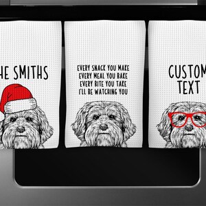 May include: Set of three white kitchen towels with a black and white illustration of a dog wearing a Santa hat, glasses, and a plain design. The text on the towels reads "THE SMITHS", "EVERY SNACK YOU MAKE EVERY MEAL YOU BAKE EVERY BITE YOU TAKE I'LL BE WATCHING YOU", and "CUSTOM TEXT".