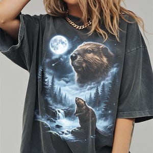 May include: Dark grey t-shirt with a detailed graphic of a beaver under a full moon, with a waterfall and forest scene. The beaver is depicted in a realistic style, with a night sky background. The shirt has short sleeves and a relaxed fit.