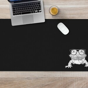 May include: Black desk mat with a white illustration of a lizard wearing sunglasses. The mat is on a wooden surface with a laptop, a coffee cup, and a computer mouse.
