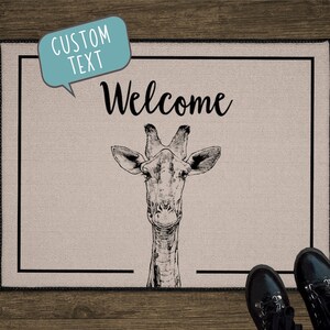 May include: A welcome mat with a black border and a beige background. The mat features a black and white illustration of a giraffe's head and the word "Welcome" in black cursive lettering. The text "Custom Text" is in a speech bubble above the giraffe.