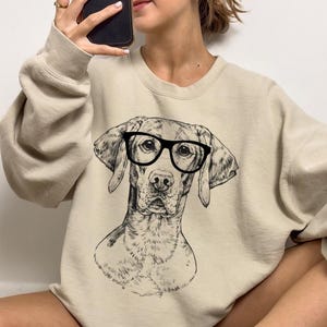May include: A beige sweatshirt featuring a black and white illustration of a dog wearing glasses. The dog is looking directly at the viewer.