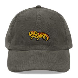 May include: A green corduroy baseball cap with an embroidered lizard design. The lizard is yellow with red spots and is smiling. The cap has a curved peak and an adjustable strap at the back. The cap is a casual accessory.
