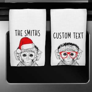 May include: Two white kitchen towels with black and white monkey illustrations. One monkey is wearing a red Santa hat and the other is wearing red glasses. The text "THE SMITHS" is printed on one towel and "CUSTOM TEXT" is printed on the other.