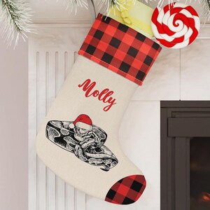 May include: A Christmas stocking with a red and black buffalo plaid cuff and heel. The stocking features a snake wearing a Santa hat and the name "Molly" in red script. A red and white swirled lollipop hangs nearby.