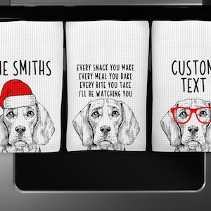 May include: Three kitchen towels with a black and white illustration of a beagle dog. The first towel has the text "THE SMITHS" and the dog is wearing a red Santa hat. The second towel has the text "EVERY SNACK YOU MAKE EVERY MEAL YOU BAKE EVERY BITE YOU TAKE I'LL BE WATCHING YOU". The third towel has the text "CUSTOM TEXT" and the dog is wearing red glasses.