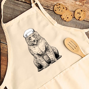 May include: Cream-colored apron featuring a detailed black and white illustration of a bear wearing a chef's hat. The apron has a pocket with a wooden spoon and is next to chocolate chip cookies. A kitchen accessory.