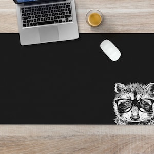 May include: Black desk mat with a white illustration of a raccoon wearing glasses. The raccoon is looking directly at the viewer.