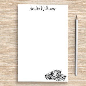 May include: White notepad with the name Amelia Williams printed at the top. The notepad has a black and white illustration of a snake at the bottom.