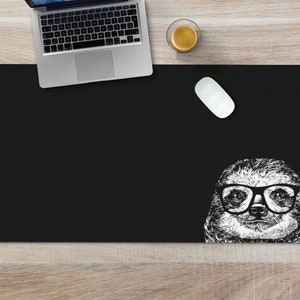 May include: Black rectangular desk mat featuring a detailed black and white illustration of a sloth wearing glasses. A laptop, a white mouse, and a cup of coffee are on a light wood surface.