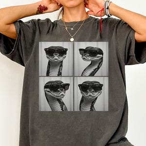 May include: Dark grey t-shirt featuring a four-panel image of a snake wearing sunglasses. The snake is in different poses in each panel. The shirt is a relaxed fit.