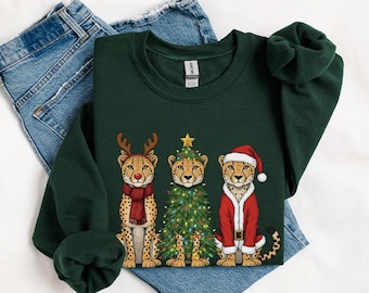 May include: Dark green sweatshirt featuring three cheetahs in festive attire. One cheetah wears reindeer antlers and a scarf, another is a Christmas tree, and the third is in a Santa suit. The sweatshirt is displayed on a pair of blue jeans.