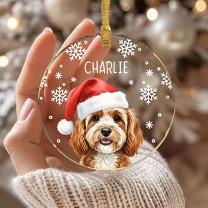 May include: A round, clear acrylic Christmas ornament featuring a printed illustration of a brown and white dog wearing a red and white Santa hat. The name CHARLIE is printed in white above the dog, surrounded by white snowflake graphics.
