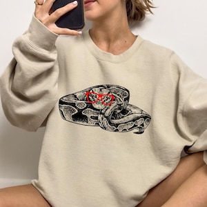 May include: A beige sweatshirt featuring a graphic of a snake wearing red glasses. The snake is curled up and has a brown and black pattern.