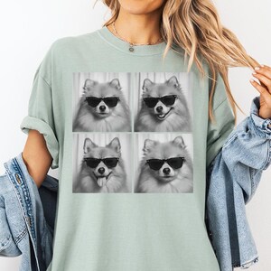 May include: A light green t-shirt featuring a black and white image of four Pomeranian dogs wearing sunglasses. The dogs have different expressions. The shirt is worn by a person wearing a denim jacket.