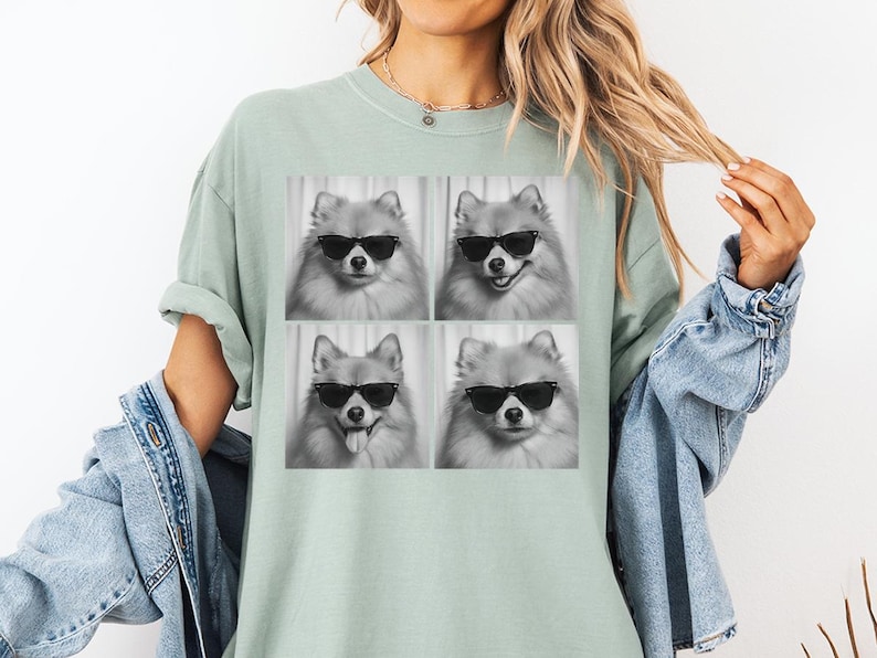 May include: A light green t-shirt featuring a black and white image of four Pomeranian dogs wearing sunglasses. The dogs have different expressions. The shirt is worn by a person wearing a denim jacket.