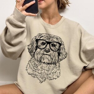 May include: A beige sweatshirt with a black and white illustration of a dog wearing glasses. The dog is a small breed with fluffy fur.