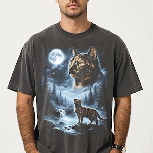 May include: Dark gray t-shirt featuring a detailed illustration of a cat in a forest scene under a full moon. The design includes a large cat face and a smaller cat standing near a waterfall, with trees and a starry sky.