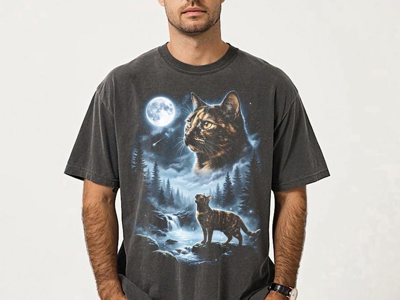 May include: Dark gray t-shirt featuring a detailed illustration of a cat in a forest scene under a full moon. The design includes a large cat face and a smaller cat standing near a waterfall, with trees and a starry sky.