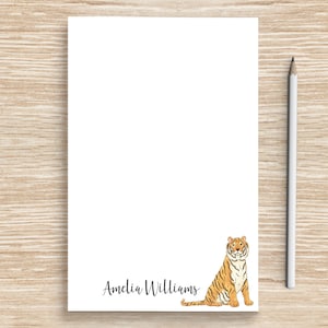 May include: A white notepad with a tiger illustration and the name "Amelia Williams" printed at the bottom. The notepad is on a woodgrain surface with a white pencil beside it.
