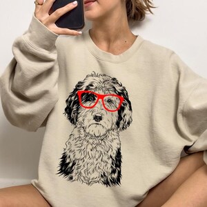 May include: A beige sweatshirt with a black and white illustration of a dog wearing red glasses. The dog's face is the central design element, with detailed fur and a serious expression. The sweatshirt has long sleeves and a crew neck.