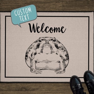 May include: A beige doormat with a black border and the word "Welcome" in black cursive lettering. A black and white illustration of a toad is centered on the mat.