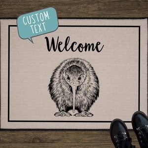 May include: A beige doormat with black trim features a black and white illustration of a kiwi bird. The text "Welcome" is written in black cursive above the bird. A speech bubble with the text "CUSTOM TEXT" is in the top left corner.
