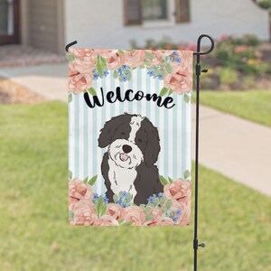 May include: A welcome garden flag with a cute black and white dog in a floral design. The flag has a blue and white striped background with the word "Welcome" in black.