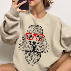 May include: A beige sweatshirt featuring a black and white illustration of a poodle wearing red glasses.