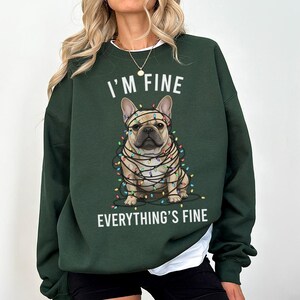 May include: Dark green sweatshirt with the text "I'M FINE EVERYTHING'S FINE". The graphic features a French Bulldog wrapped in Christmas lights. The sweatshirt has long sleeves and a crew neck.
