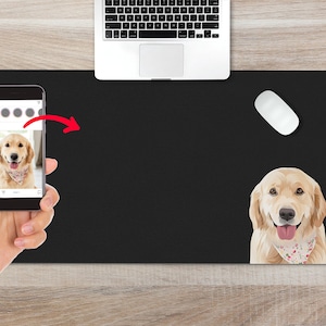 May include: A black desk mat with a white mouse and a golden retriever dog illustration. The dog is wearing a pink and white floral bandana. A hand is holding a smartphone with a photo of a golden retriever dog on the screen.