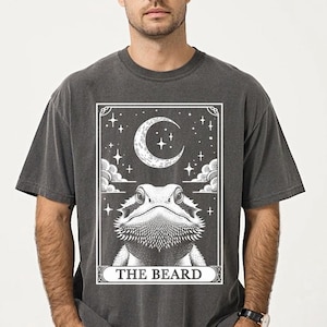 May include: A dark gray t-shirt with a white graphic of a bearded dragon in a tarot card style design. The card reads "The Beard" and features a crescent moon and stars.