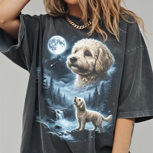 May include: Dark gray t-shirt featuring a detailed illustration of a dog under a full moon. The design includes a waterfall, forest, and a starry sky. The dog is depicted in two forms, one looking up at the moon and the other as a portrait.